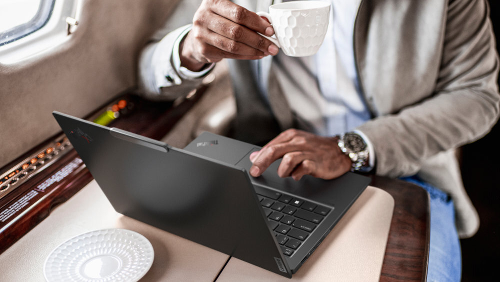Mężczyzna w eleganckim ubraniu pracuje na laptopie Lenovo ThinkPad X1 w luksusowym wnętrzu przypominającym prywatny odrzutowiec, trzymając filiżankę kawy; czarna, smukła obudowa laptopa podkreśla mobilność i profesjonalny charakter urządzenia.