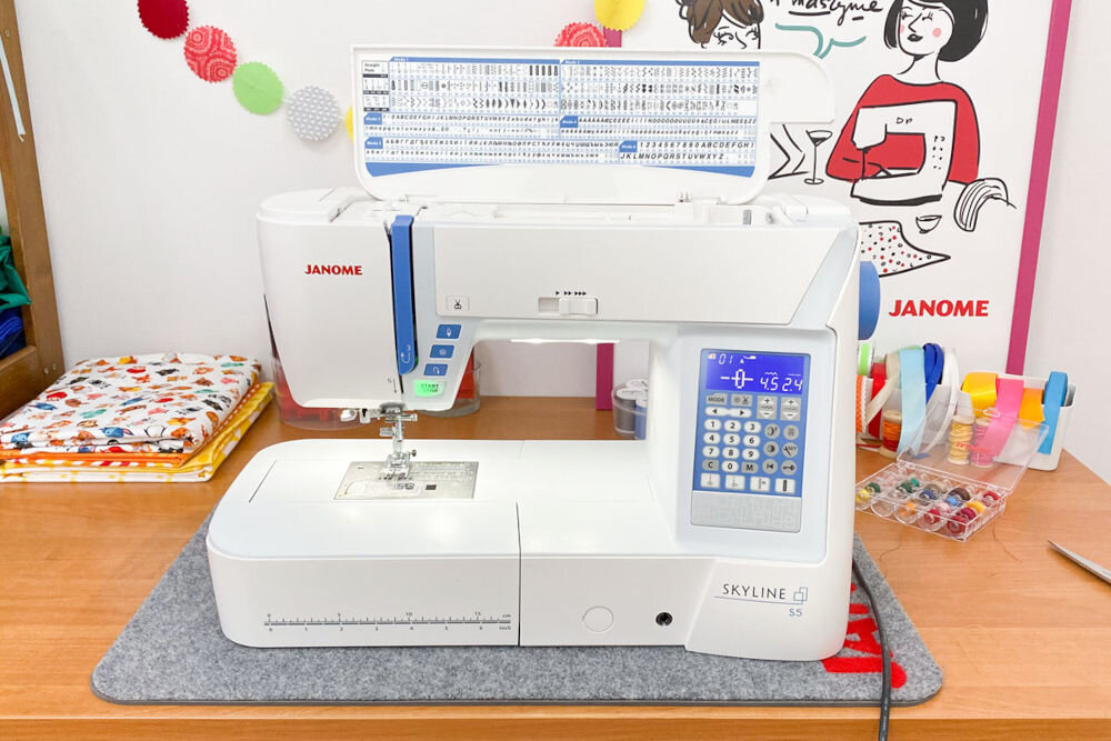 Maszyna do szycia JANOME Skyline S5 maszyna stoi na blacie niebieskie elementy ekran przyciski suwaki narysowane ściegi dla pasjonatów szycia