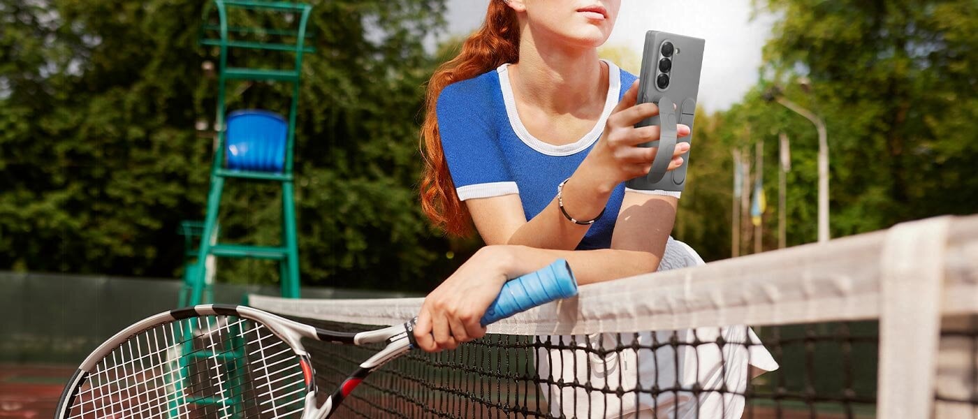 Młoda kobieta na korcie tenisowym opiera się o siatkę kortu tenisowego, trzymając wygodnie Galaxy Z Fold6. Telefon znajduje się w szarym etui Silicone Case, a kobieta trzyma telefon z uchwytem etui przeplecionym przez jej palce. 